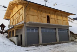 Alpines Gebäude mit Holzfassade im oberen Bereich und Beton im Erdgeschoss, ausgestattet mit drei nebeneinanderliegenden grauen Garagentoren