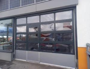 Deckensektionaltor mit Eingangstür, gefertigt aus dunklem Rahmen und mehreren transparenten Fenstern.