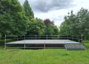 Freistehendes Bühnengerüst auf einer Wiese, mit Geländer und einer Treppe an der rechten Seite umgeben von Bäumen